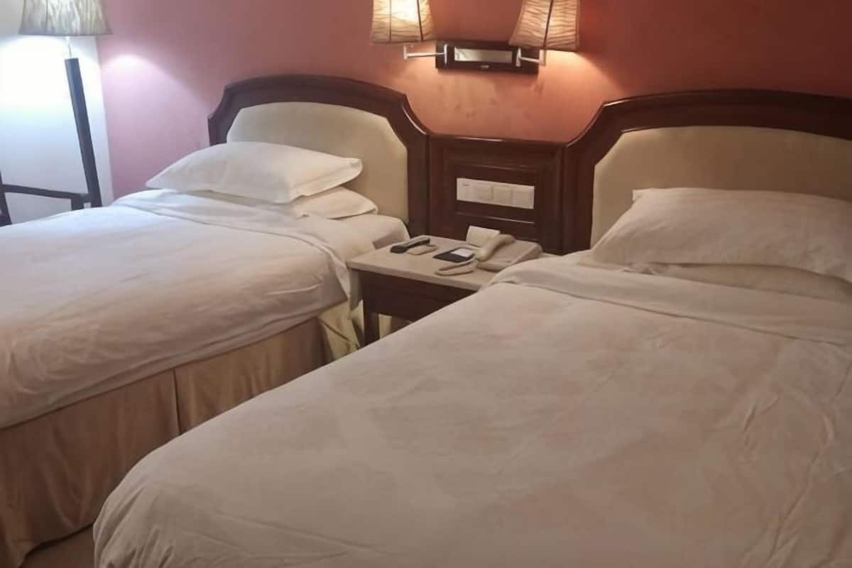 Quarto de hotel com duas camas, uma mesa de cabeceira com telefone e óculos, e uma luminária na parede.