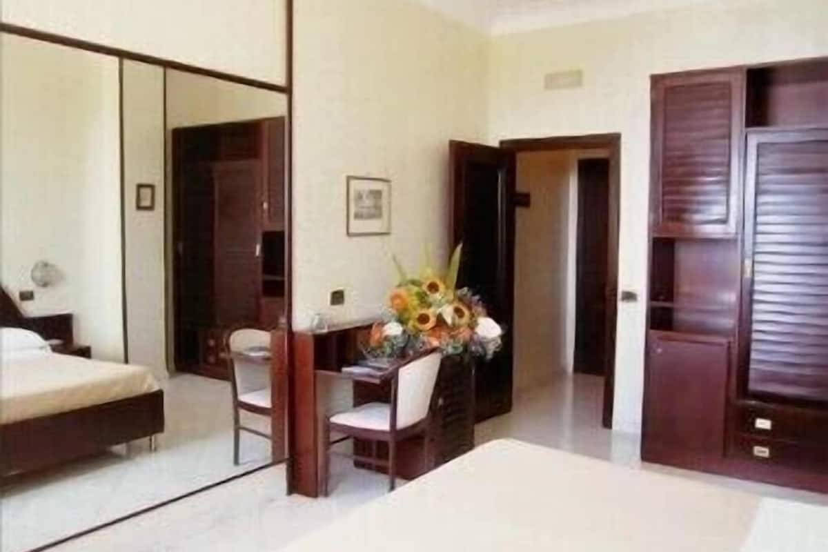 A hotel room with a bed, a desk, a chair, a mirror, and a door.