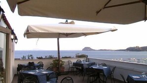 Terraza al aire libre con mesas y sillas, protegida por sombrillas, con vistas a la costa y barcos.