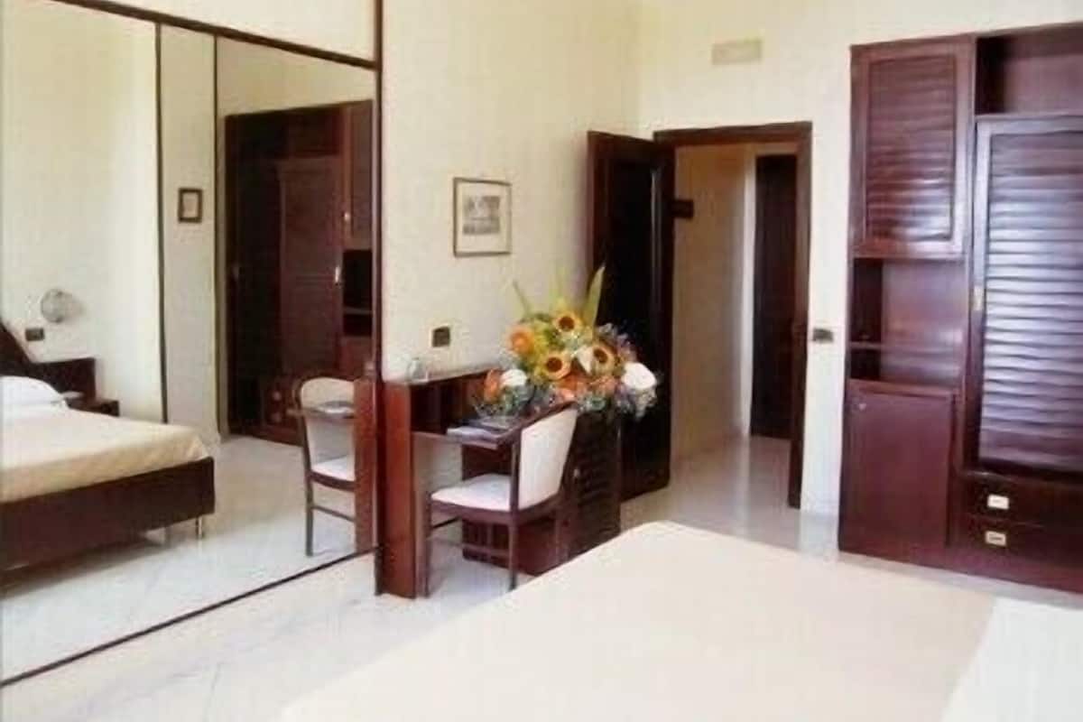 A hotel room with a bed, a desk with a chair, a mirror, and a wooden wardrobe.