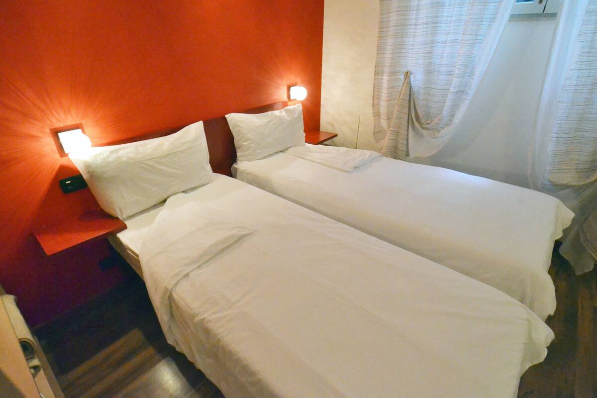A hotel room with two single beds, a wooden floor, a red accent wall, and a window with curtains.