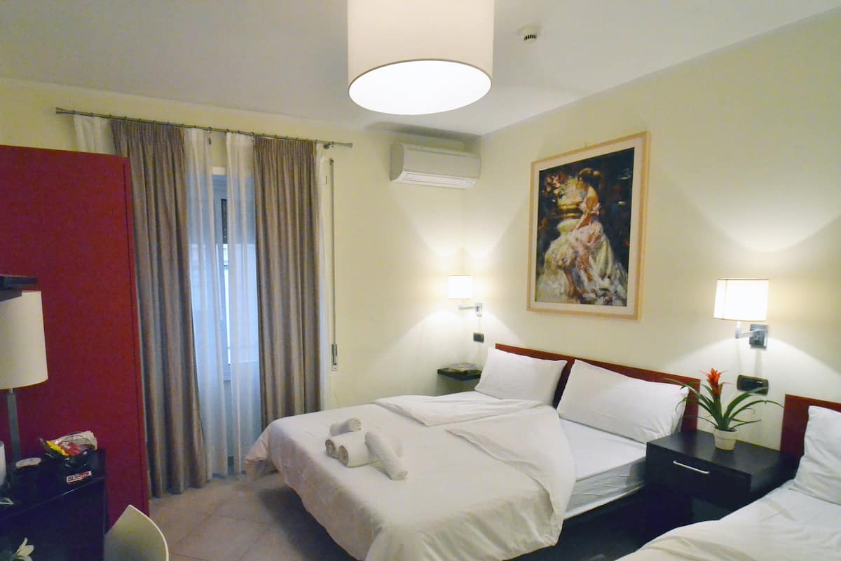 A hotel room with two beds, a desk, a chair, a painting on the wall, and an air conditioning unit.