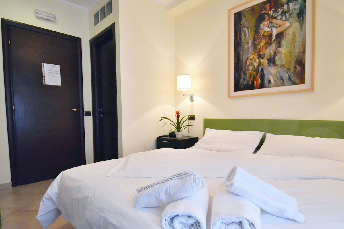 A hotel room with a neatly made bed, a nightstand with a lamp and a plant, and a painting on the wall.