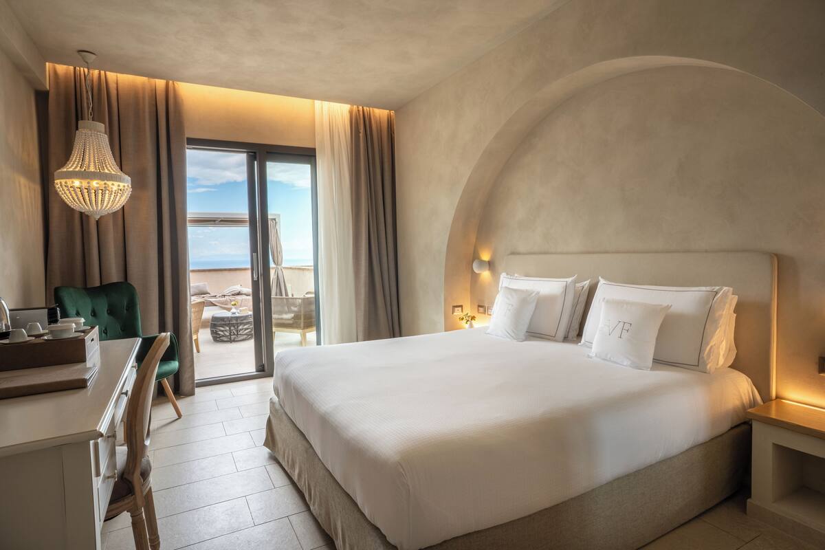 Une chambre d’hôtel avec un grand lit, un bureau, une chaise et une vue sur la mer, accessible par une porte coulissante.