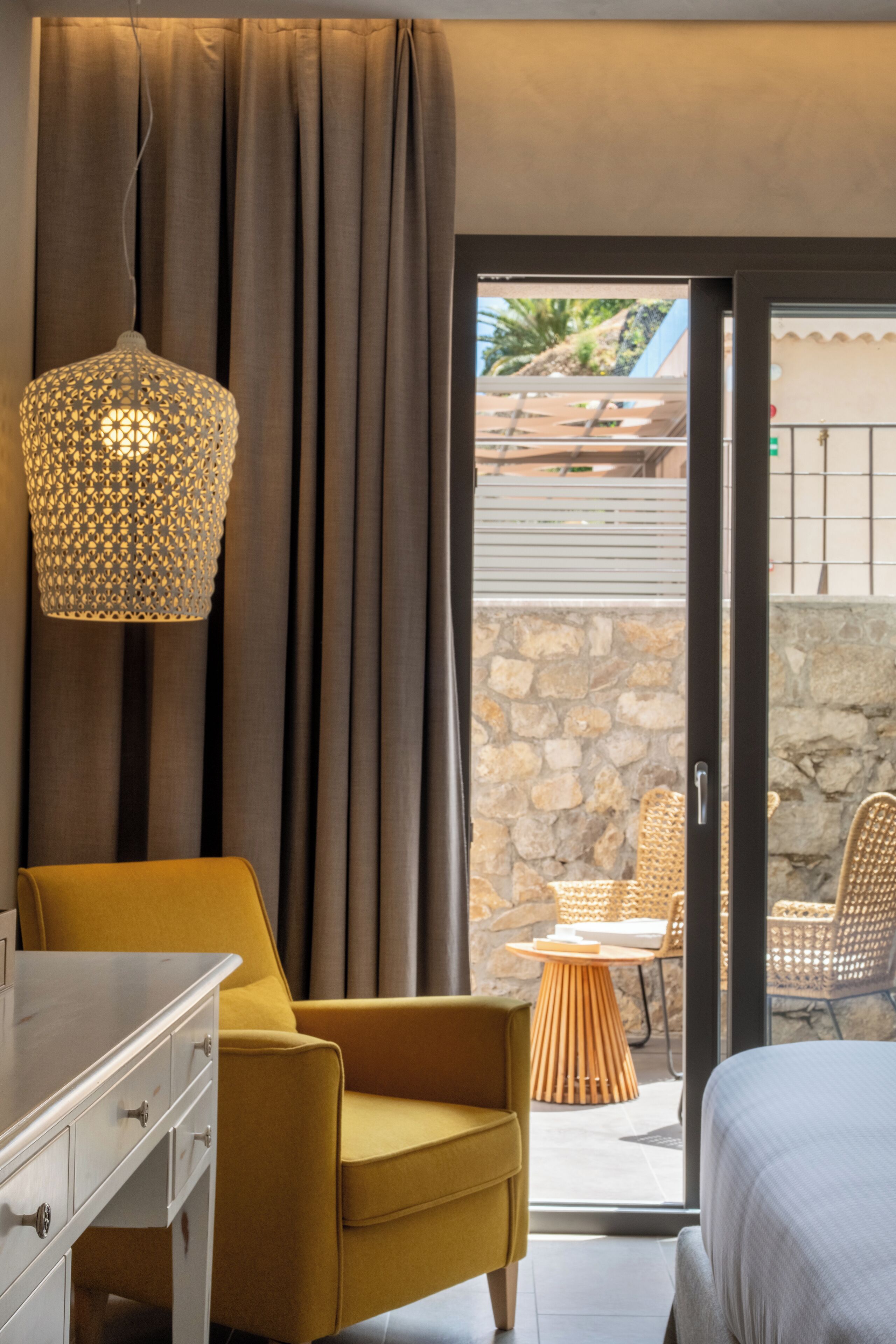 A hotel room with a bed, a yellow armchair, a white dresser, and a wicker chair outside.