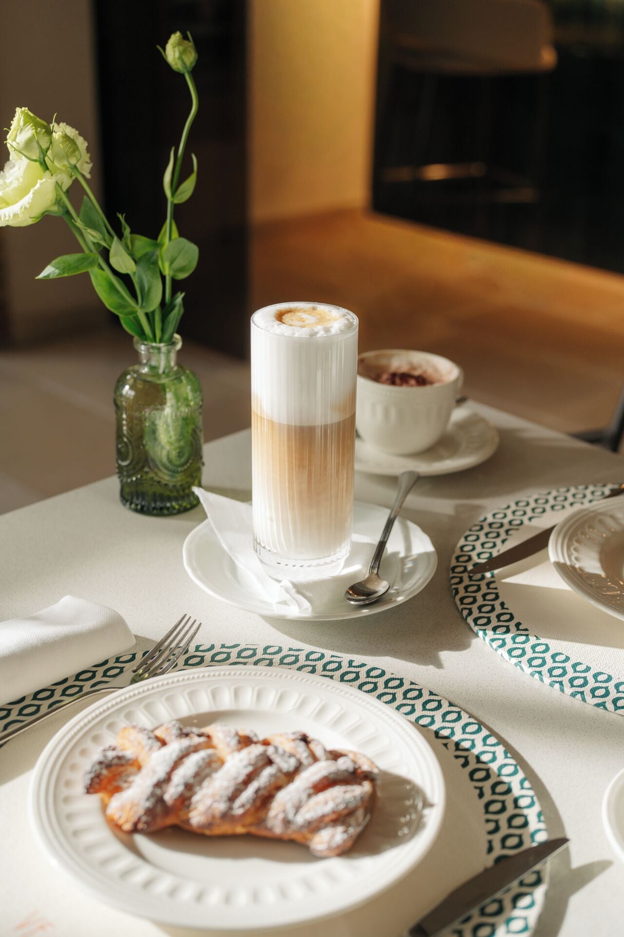 A table set with a tall glass of layered beverage, a plate with a pastry, a small vase with flowers, and a bowl of dessert.