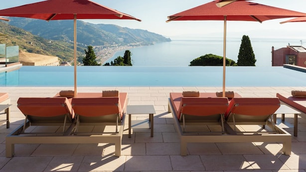 A poolside area with red umbrellas, lounge chairs, and a view of the sea and hills. - Villa Fiorita Boutique Hotel (Taormina)