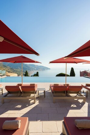 Ein Poolbereich mit roten Sonnenschirmen, Liegestühlen und Blick auf das Meer und die Hügel.