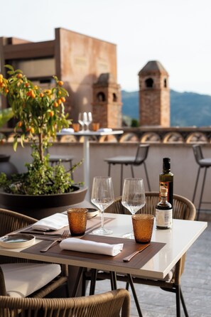Ein Esstisch ist gedeckt mit Weingläsern, einer Flasche und einem Blumentopf auf einer Terrasses mit Korbstühlen und Blick auf ein historisches Gebäude im Hintergrund.