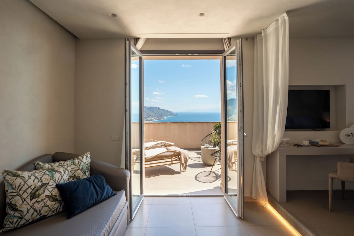 Une chambre moderne avec un balcon, un canapé agrémenté de coussins à motifs, un bureau avec une télévision et une vue sur la mer.