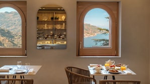 A dining area with a view of a coastal landscape through arched windows, a buffet with dishes, and breakfast items on the table. - Villa Fiorita Boutique Hotel (Taormina)