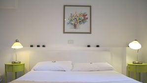 Un lit bien fait, recouvert de draps blancs, flanqué de deux tables de chevet vertes sur lesquelles sont posées des lampes, et une œuvre d’art florale encadrée au-dessus de la tête de lit.