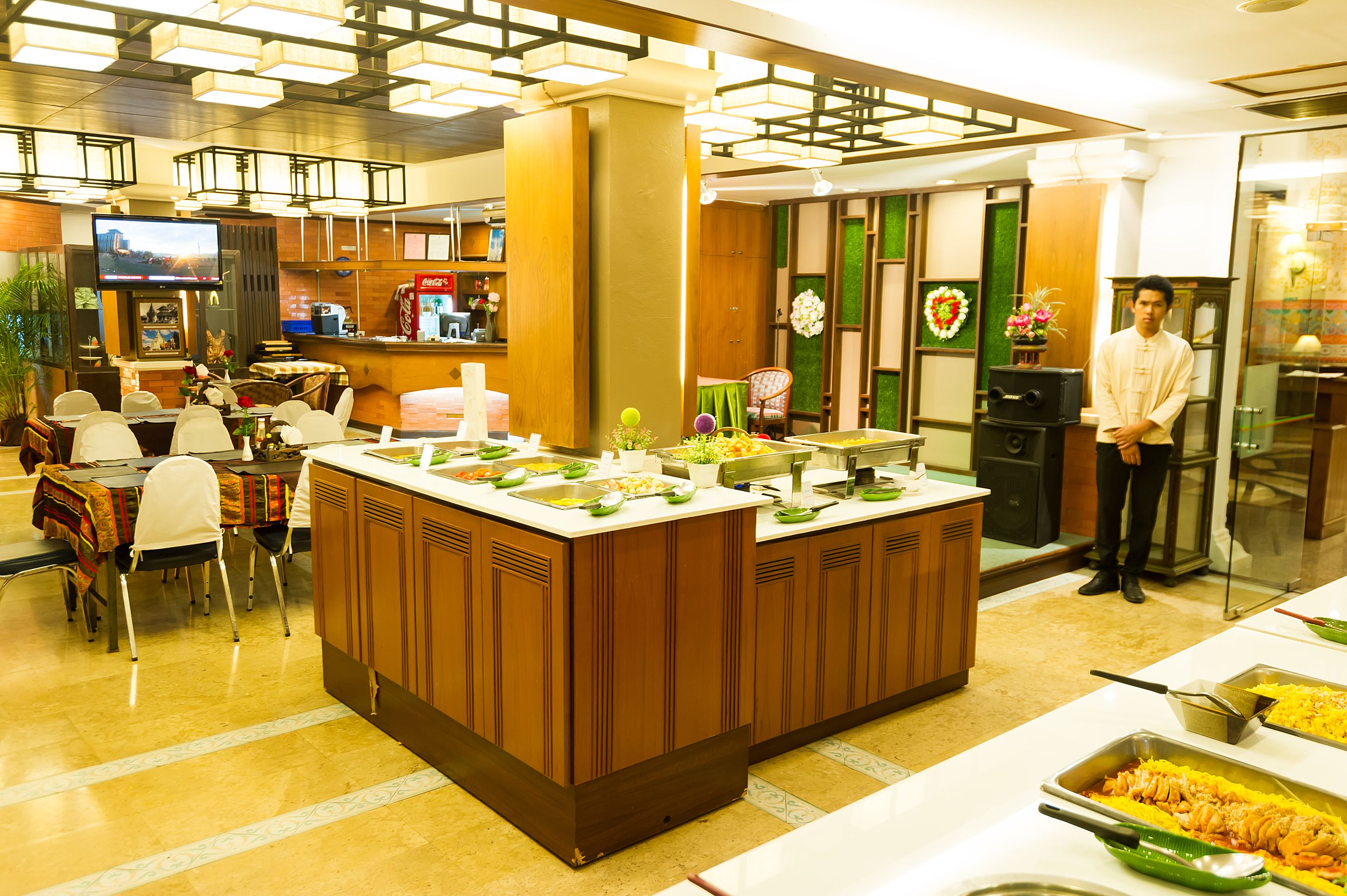 A hotel lobby with a central island featuring a buffet, seating area with chairs, a television, and a Coca-Cola refrigerator.