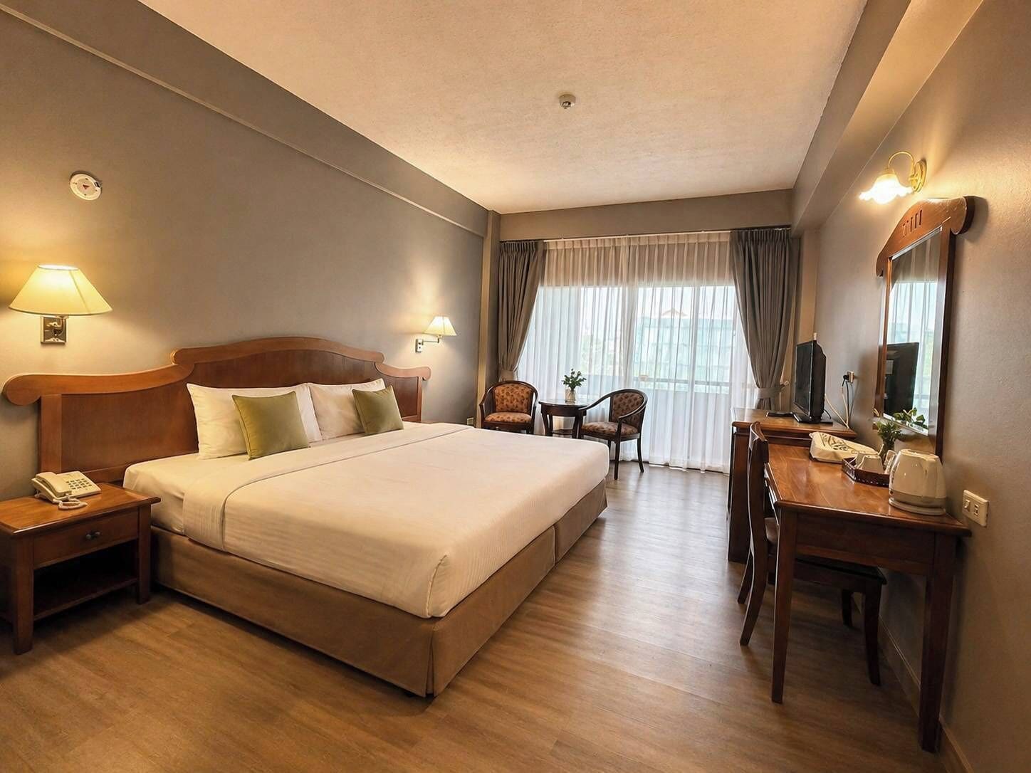 A hotel room with a large bed, bedside lamps, a desk with a chair, a small table with a chair, and a window with curtains.