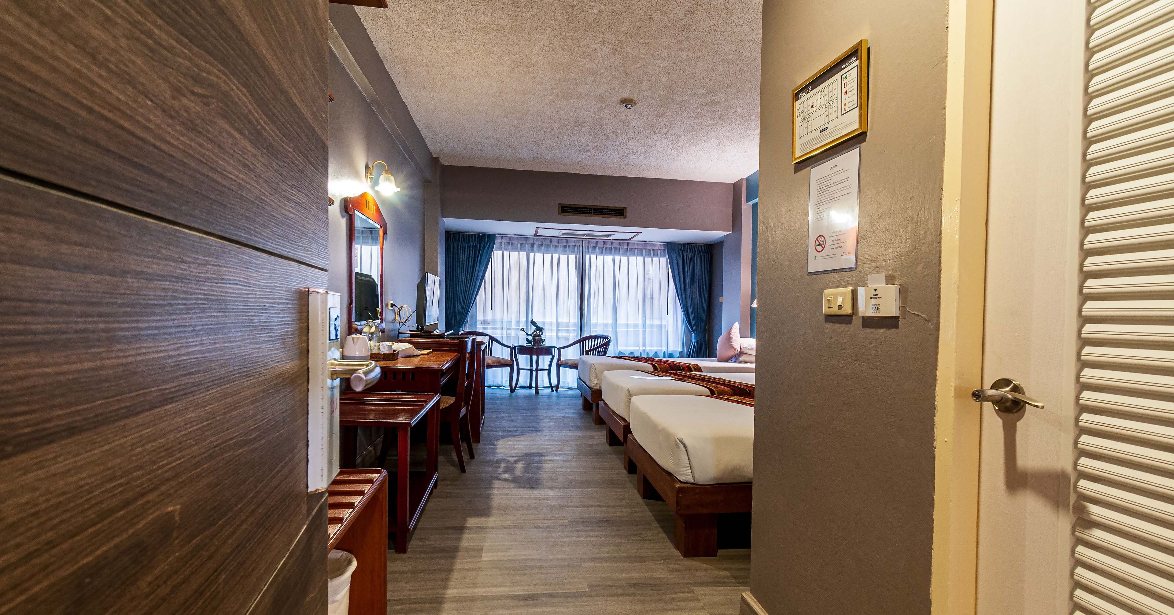 Ett hotellrum med en garderob i trä, ett skrivbord med en stol, ett litet bord med en stol, två sängar, ett fönster med gardiner och en dörr med en ljusströmbrytare.