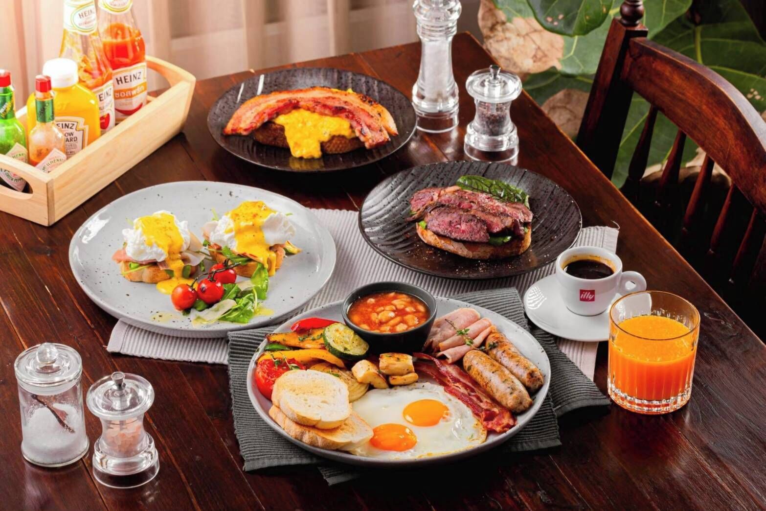 A breakfast setting with plates of eggs, bacon, sausages, and toast, along with condiments and a glass of orange juice.