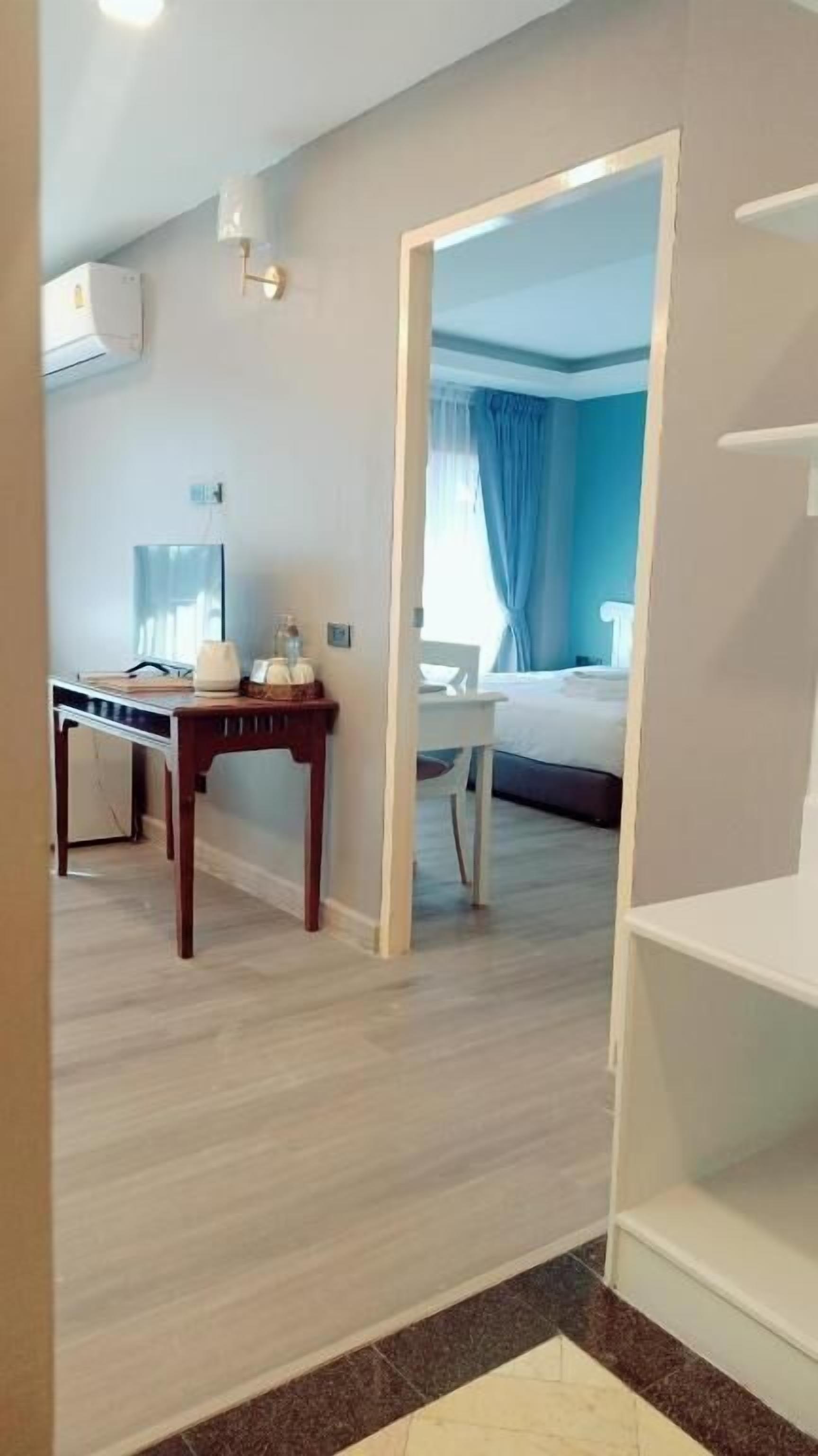 A hotel room with a bed, a desk, and a chair. There is a window with curtains, and a white shelf unit.