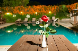 A wooden table with a white vase containing a single red rose, positioned in front of a swimming pool.