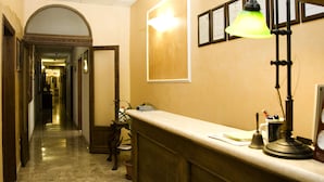 A hotel reception area with a marble floor, a reception desk, a lamp, and framed certificates on the wall. - Hotel Merlini (Florence)