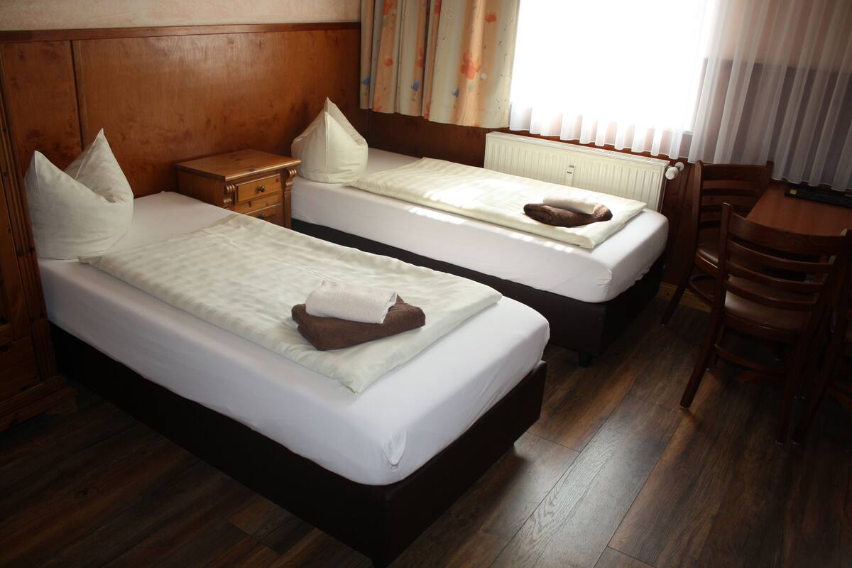 Una habitación de hotel con dos camas individuales, cabeceros y mesitas de noche de madera, una mesa con sillas, y una ventana con cortinas.