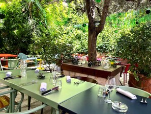 An outdoor dining area with green tables and chairs, surrounded by lush greenery and trees. - Saint Louis (Aigues-Mortes)