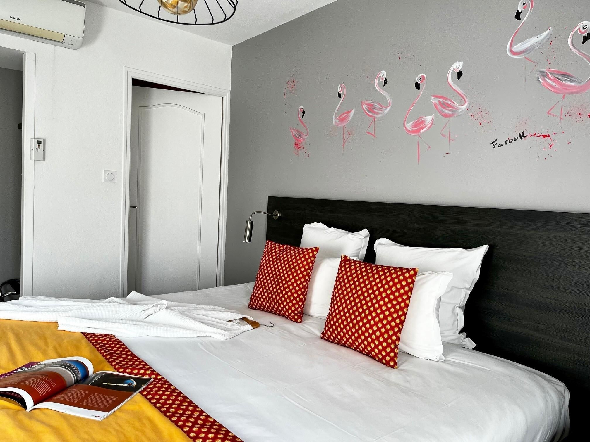 Una camera d'albergo con un letto, biancheria da letto bianca, cuscini rossi con motivo decorativo, un murale con fenicotteri e una porta.