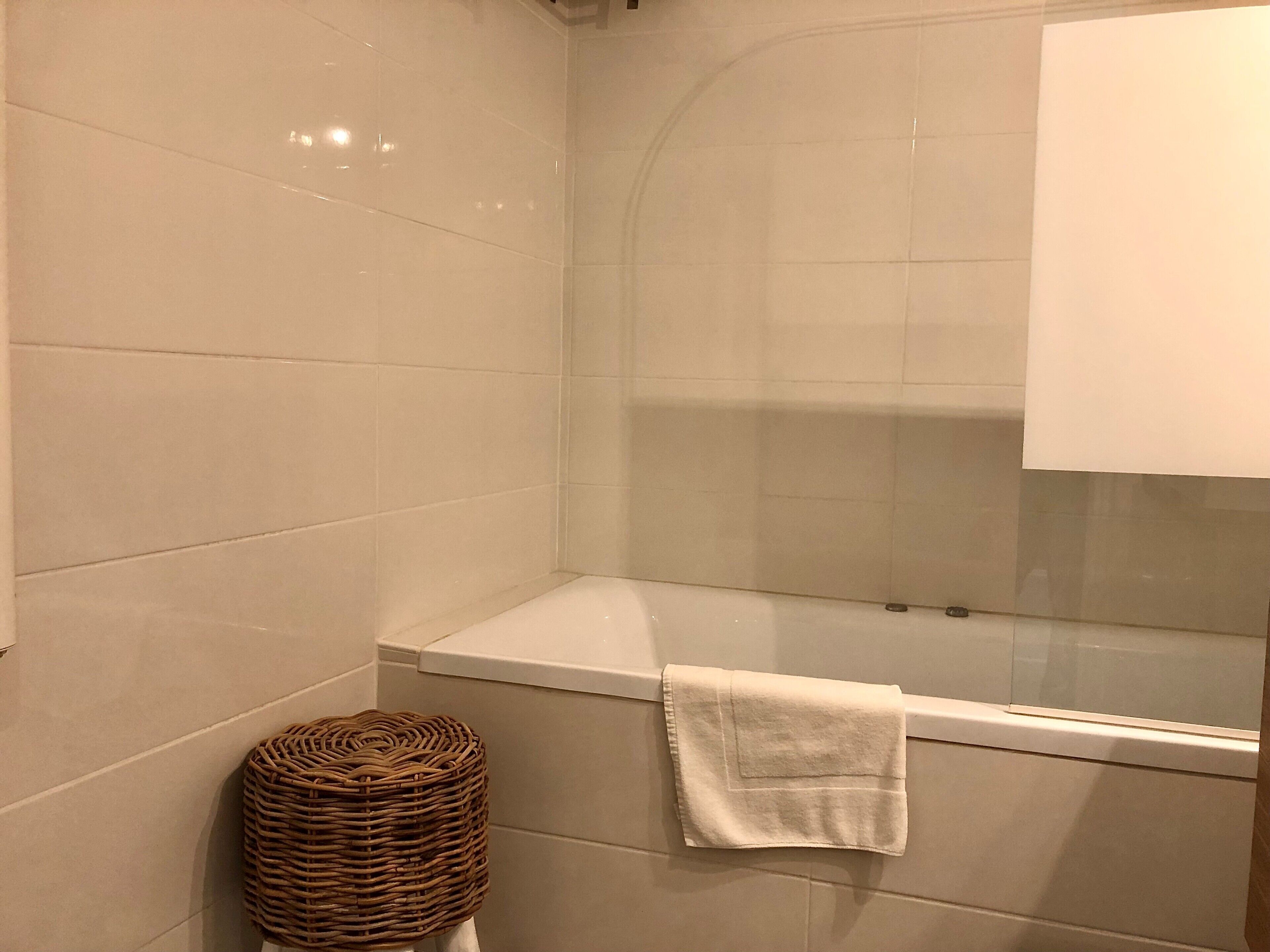 Un baño con baldosas blancas, una bañera, una cesta de mimbre y una toalla colgada en la pared.