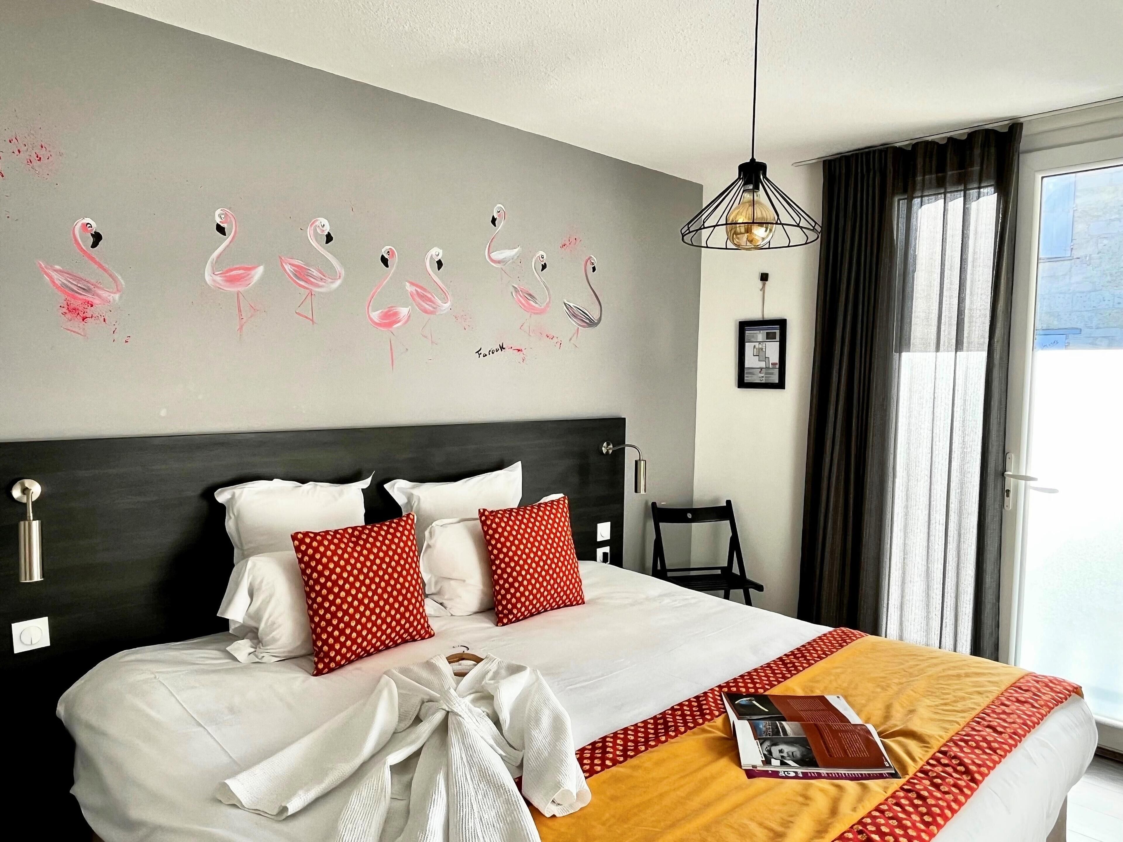 Una habitación de hotel con una cama, almohadas rojas y blancas, un plaid amarillo, una lámpara colgante y un mural de flamencos en la pared.
