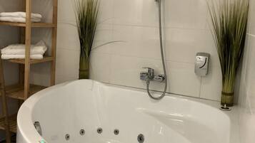 A bathroom with a large white bathtub, a wooden shelf with folded towels, and two tall plants.