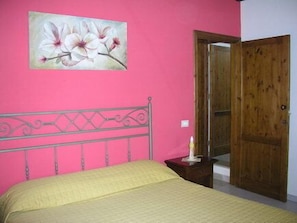 Een slaapkamer met een roze muur, een bed met een gele sprei, een houten deur en een schilderij van bloemen.