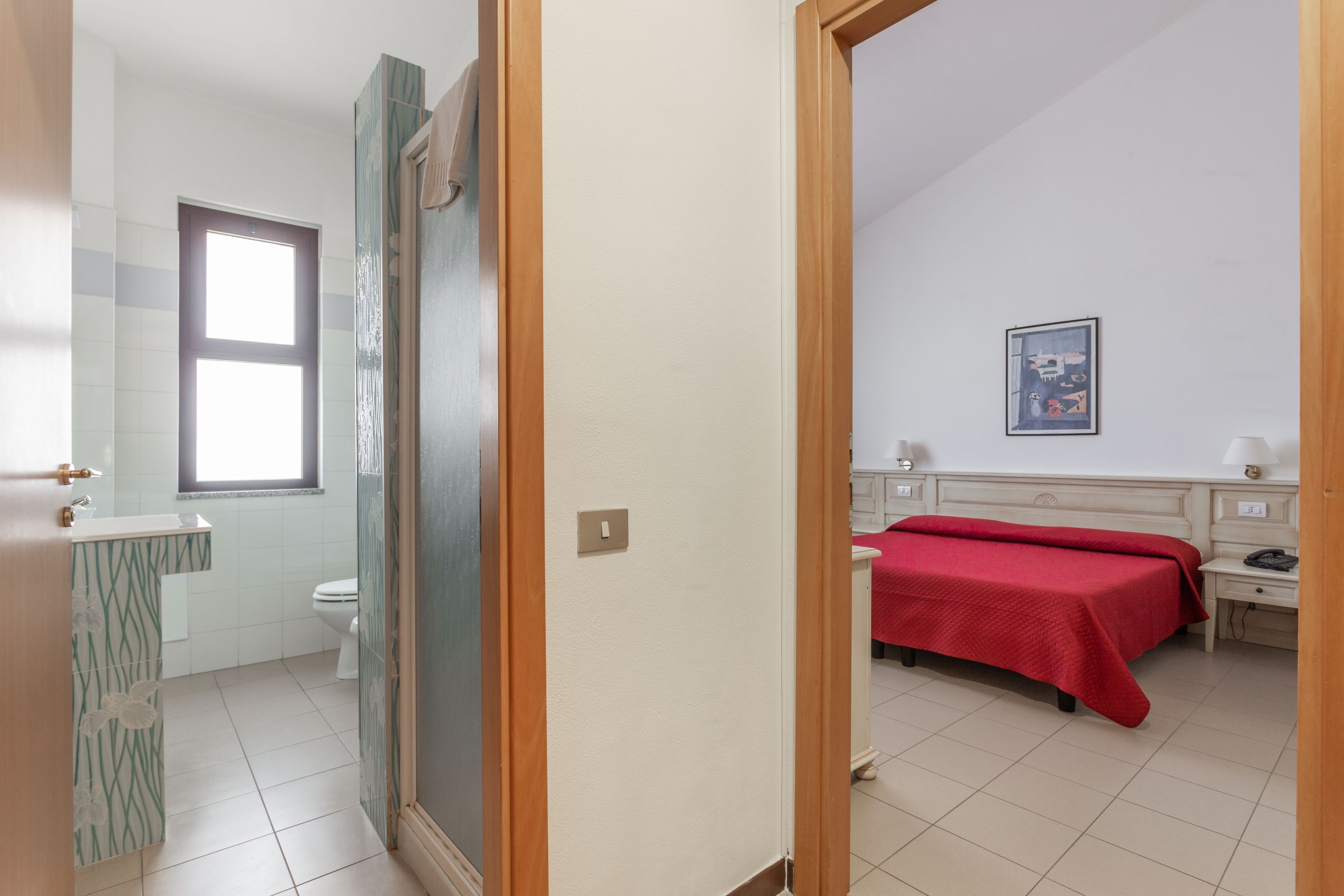 A bathroom with a toilet, sink, and a window. There is a bed with a red bedspread in the adjacent room.