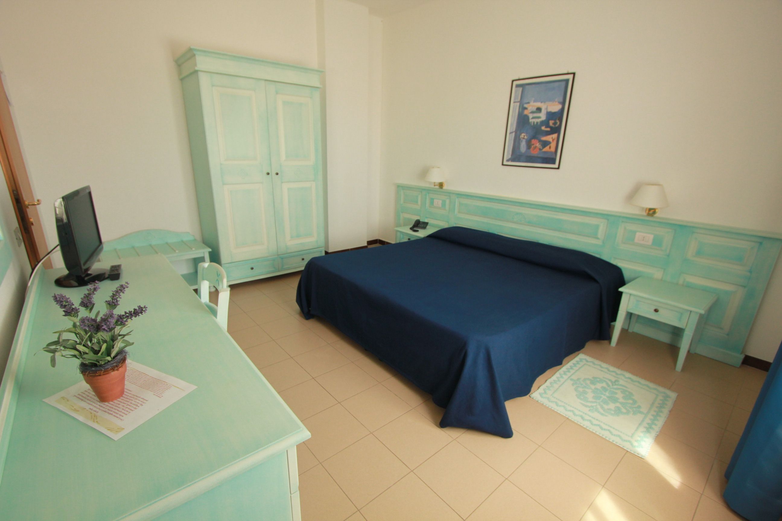 A room with a bed, a desk with a computer, a wardrobe, and a small table with a lamp.