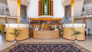A hotel lobby with a reception desk, two potted plants, a large patterned carpet, and a central column with vertical stripes.