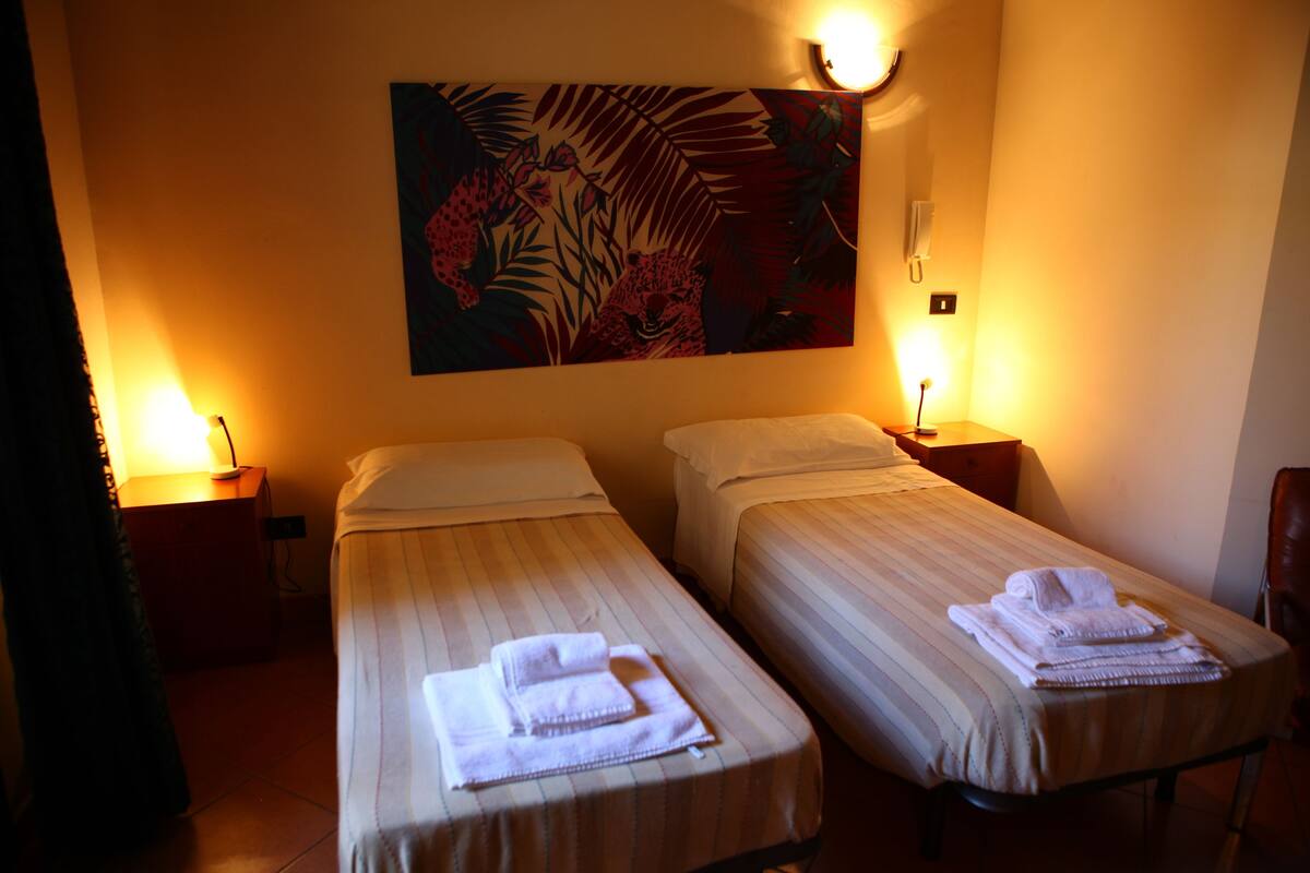 Una camera d'albergo con due letti singoli, ciascuno con un asciugamano piegato e un comodino con una lampada.