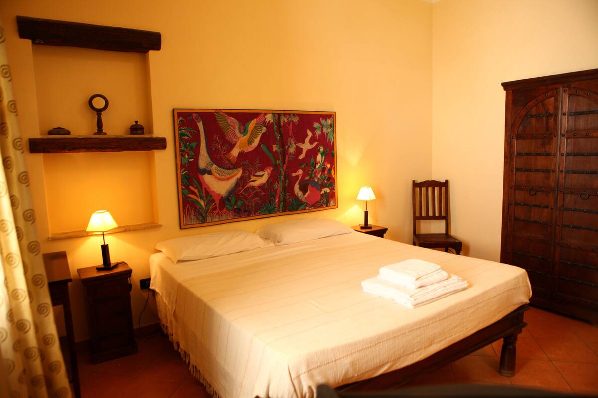Una camera d'hotel con un letto, due lampade ai lati, un armadio in legno, un quadro appeso al muro e uno scaffale con una chiave e un piccolo contenitore.