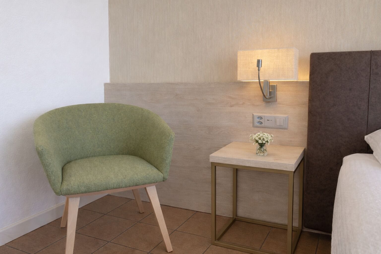 Ein Hotelzimmer mit einem grünen Sessel, einem kleinen Nachttisch mit einer Vase und einer Wandlampe.