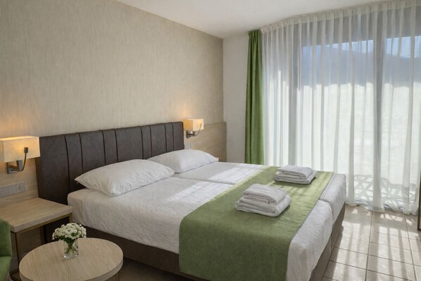 A hotel room with a bed, two bedside lamps, a round table with a vase of flowers, and a view of the outdoors through sheer curtains.