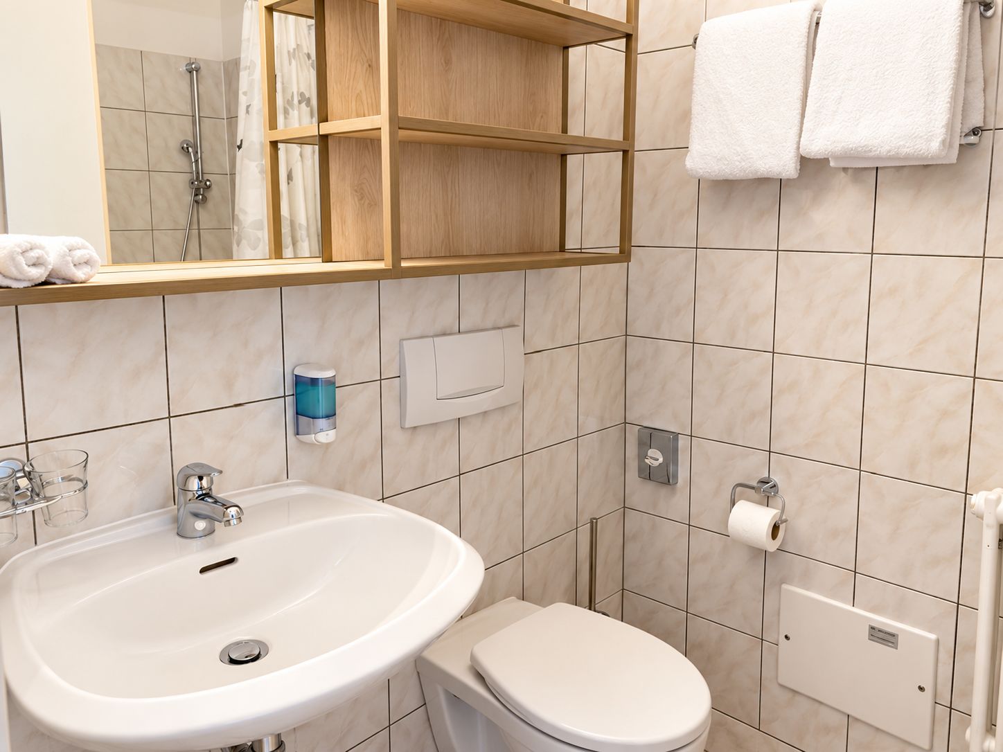 Un baño con lavamanos blanco, inodoro, ducha y espejo con repisa de madera encima.
