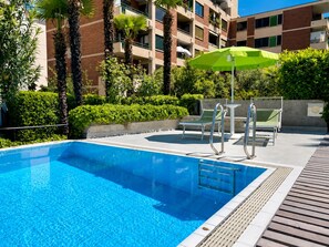 A swimming pool with a clear blue water, surrounded by a wooden deck, and a green umbrella. - Hotel Nessi Locarno (Locarno)