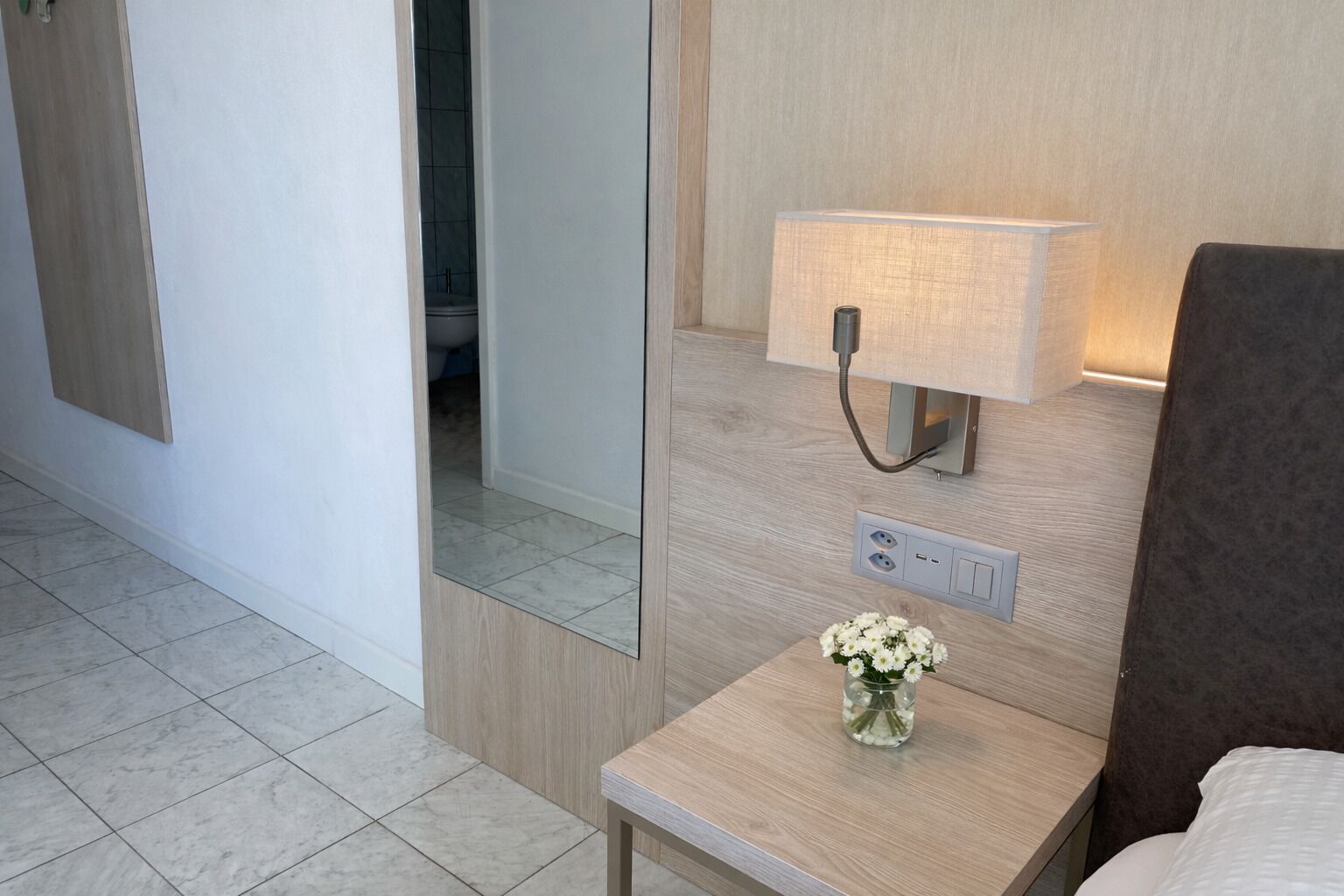 A hotel room with a bedside table, a lamp, a mirror, and a vase with flowers.