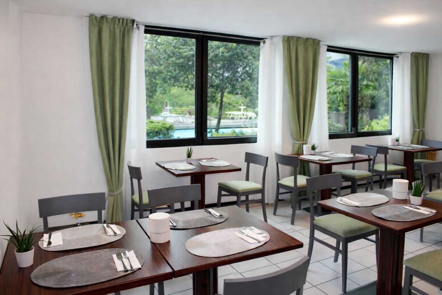 A restaurant with multiple tables and chairs, large windows with curtains, and a view of greenery outside.