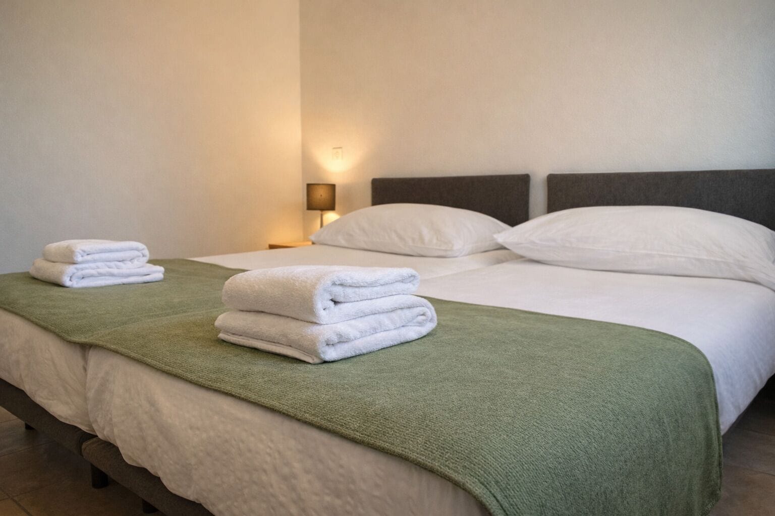 Une chambre d’hôtel avec un lit, deux serviettes blanches, un plaid vert et une table de chevet avec une lampe.
