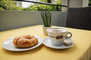 Una mesa con un croissant, una taza de café con un logo, una pequeña planta en maceta y vistas a la vegetación a través de una ventana.