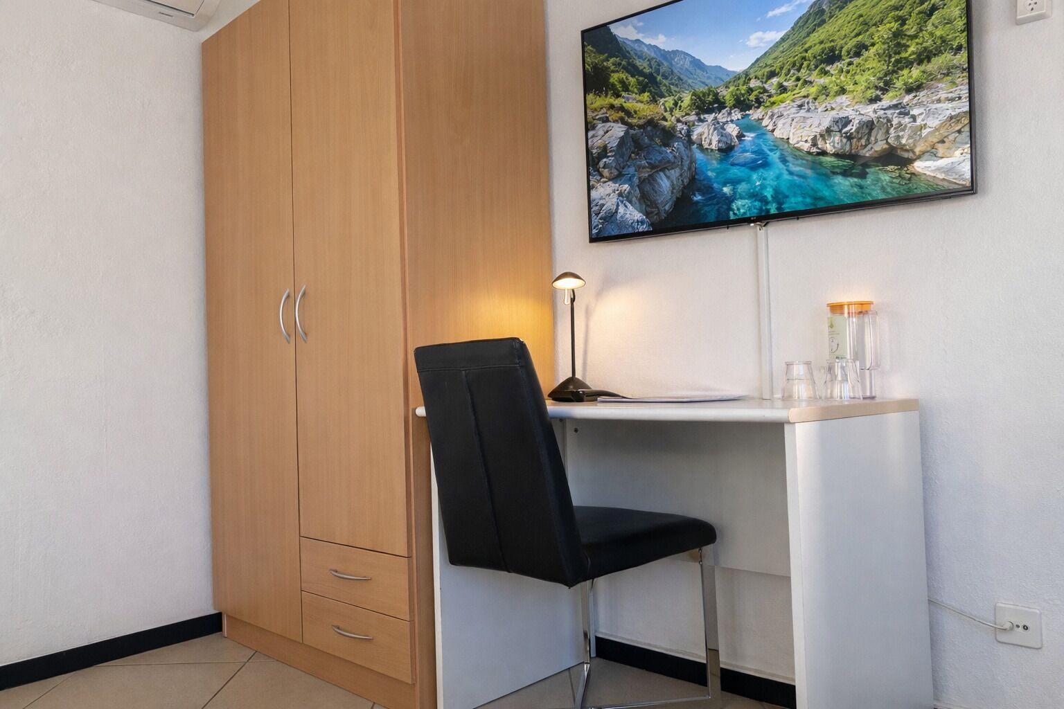 A room with a wooden wardrobe, a desk with a chair, a desk lamp, and a framed picture of a mountainous landscape.