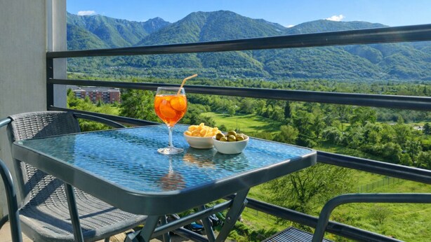 Ein Balkon mit Tisch, gedeckt für eine Mahlzeit, einem Getränk und Blick auf Berge.