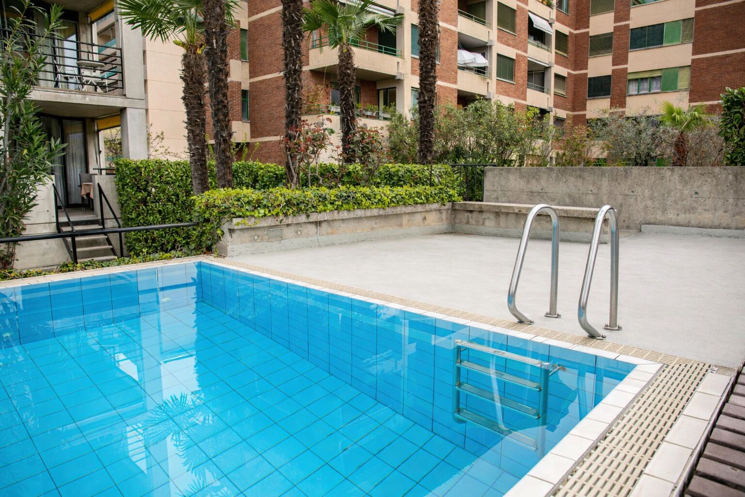Une piscine avec un bord carrelé, un escalier en métal et une terrasse en béton.