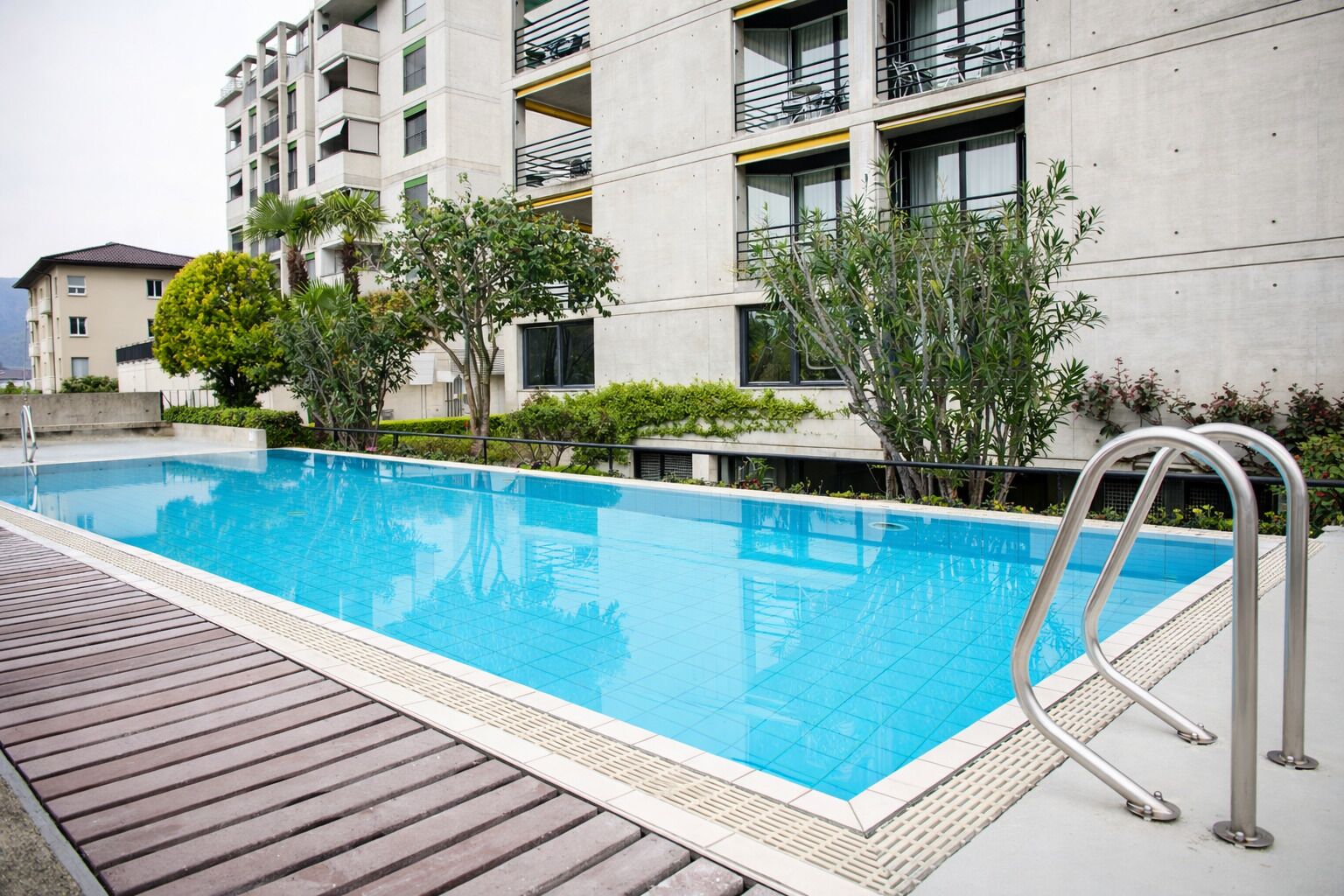 Piscina rectangular con muelle de madera, rodeada de vegetación y edificios modernos.
