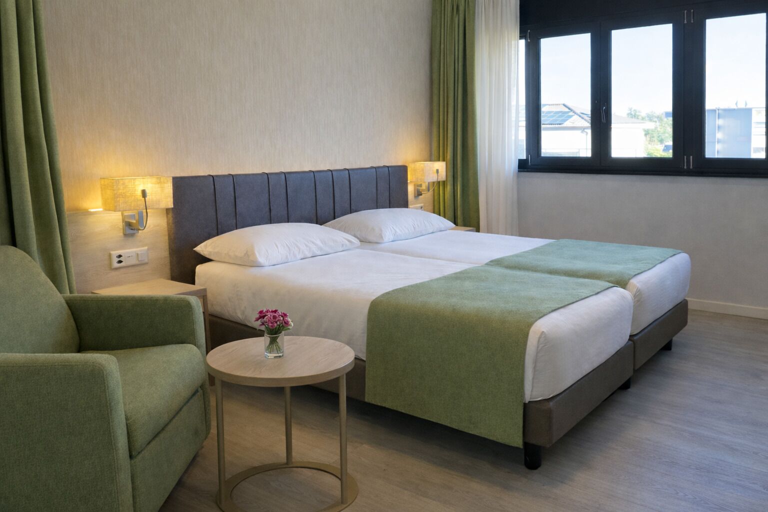 Une chambre d’hôtel avec un lit double, un fauteuil vert, une petite table ronde ornée d’un vase de fleurs et une vue sur l’extérieur par une fenêtre.