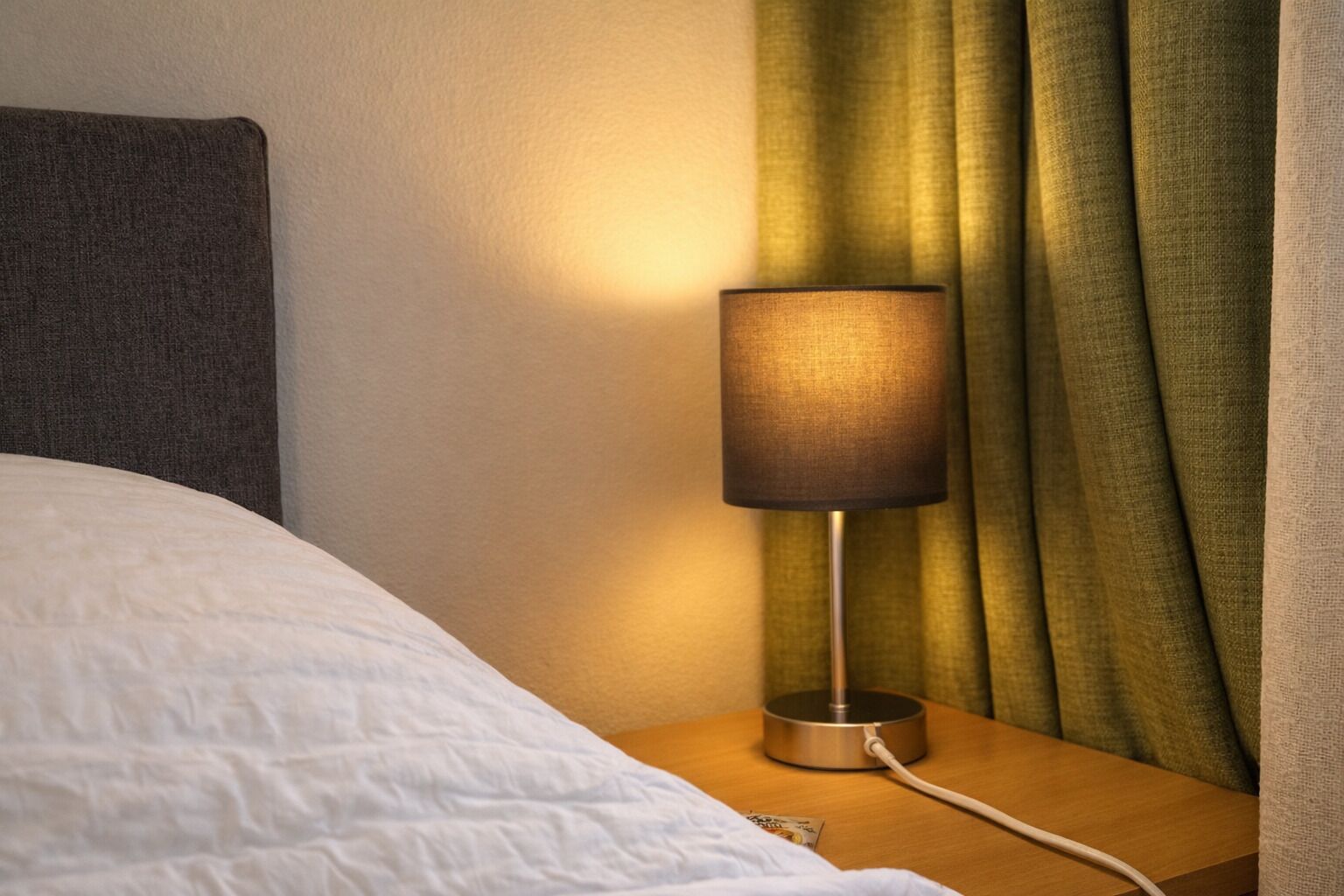 Un lit avec une couette blanche et une table de chevet sur laquelle se trouvent une lampe et un câble d’alimentation.