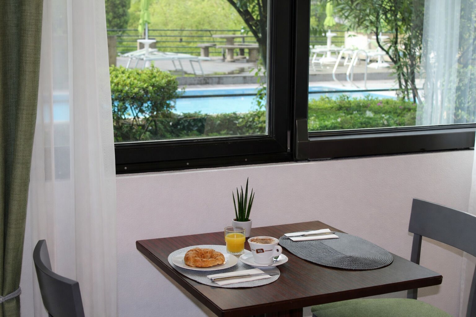 Una mesa de desayuno con un croissant, una taza de café y un vaso de zumo de naranja, frente a una ventana con vistas a una piscina y follaje.